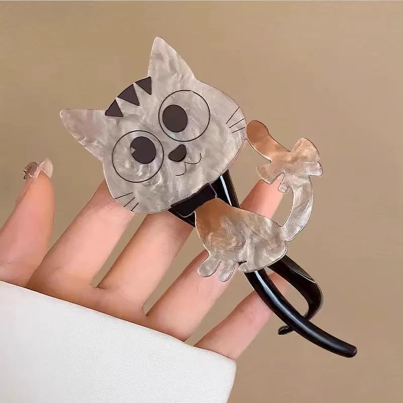 A close-up of a hand holding a black hair claw clip decorated with two translucent, cat-shaped acrylic pieces in a light gray and beige marbled pattern, against a solid beige background.