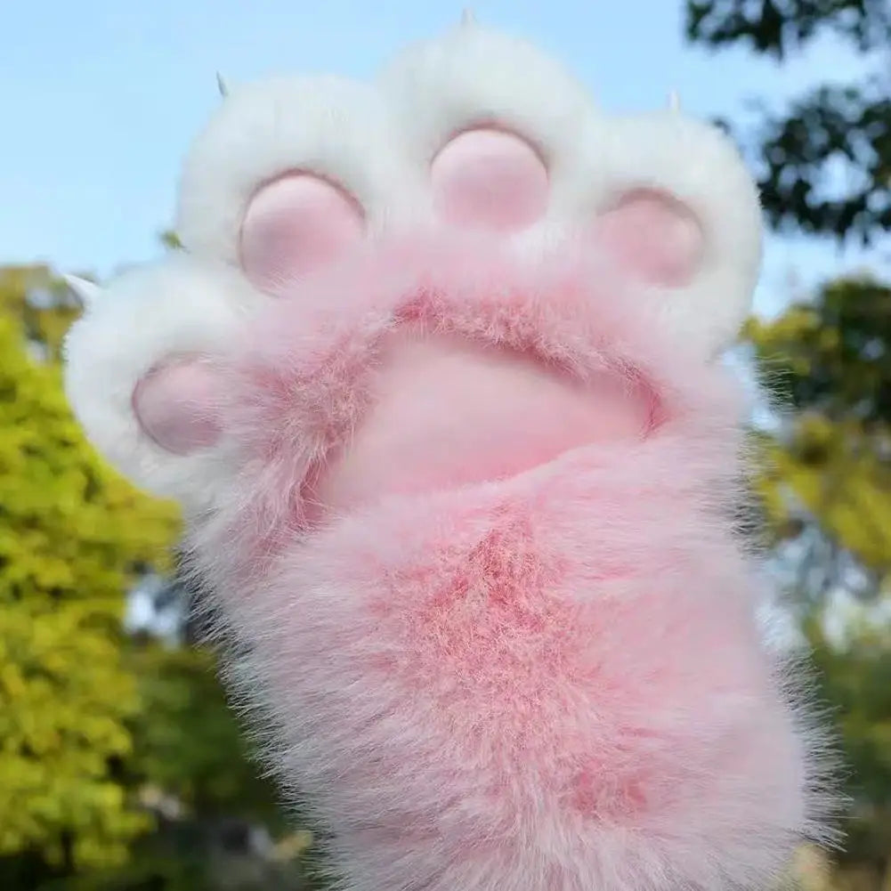A fluffy faux-fur paw-shaped accessory, likely a wrist warmer or glove, featuring a white outer paw with pink pads and a soft pink inner body, shown against a blurred outdoor background of green trees and blue sky.