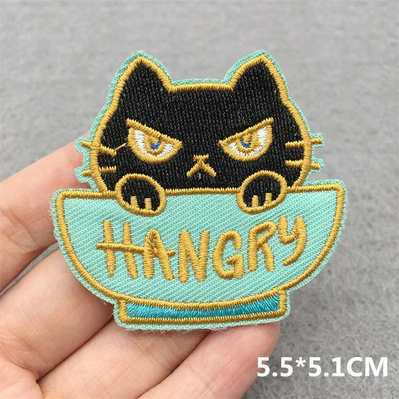 Embroidered patch of a black cat in a bowl with 'HANGRY' text held by a hand on a gray background.