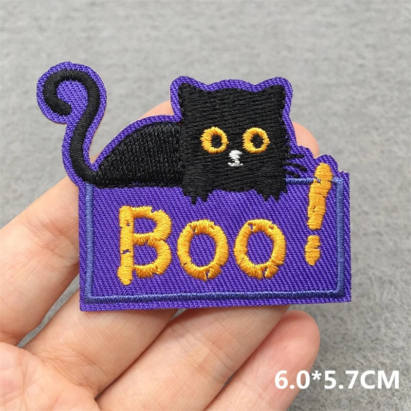 Halloween-themed patch with a black cat and 'BOO!' text held by a hand on a gray background.