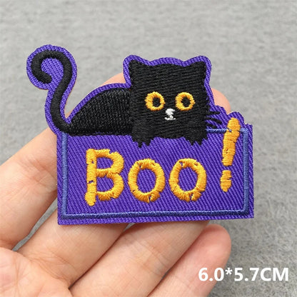 Halloween-themed patch with a black cat and 'BOO!' text held by a hand on a gray background.