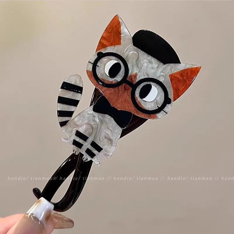 Cat-shaped hair clip with glasses and striped tail held by a hand against a neutral background