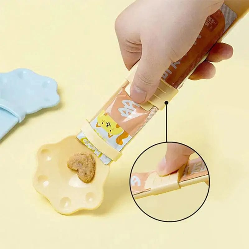 A hand using the Cat Strip Squeezer Treat Spoon to dispense a cat treat from a pouch onto a spoon, with a close-up inset showing the treat being served to a cat.