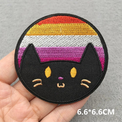 Embroidered patch of a black cat face with rainbow stripes held by a hand on a gray background.