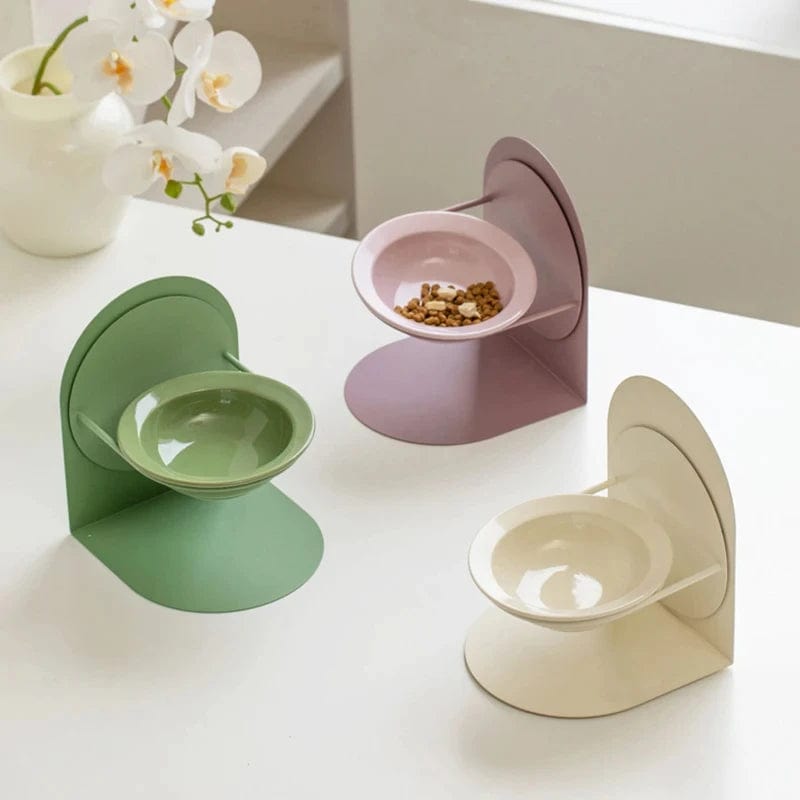 Three ceramic cat bowls in green, pink, and white colors, each with a adjustable stand, displayed on a table with a white cat food bowl in the background.