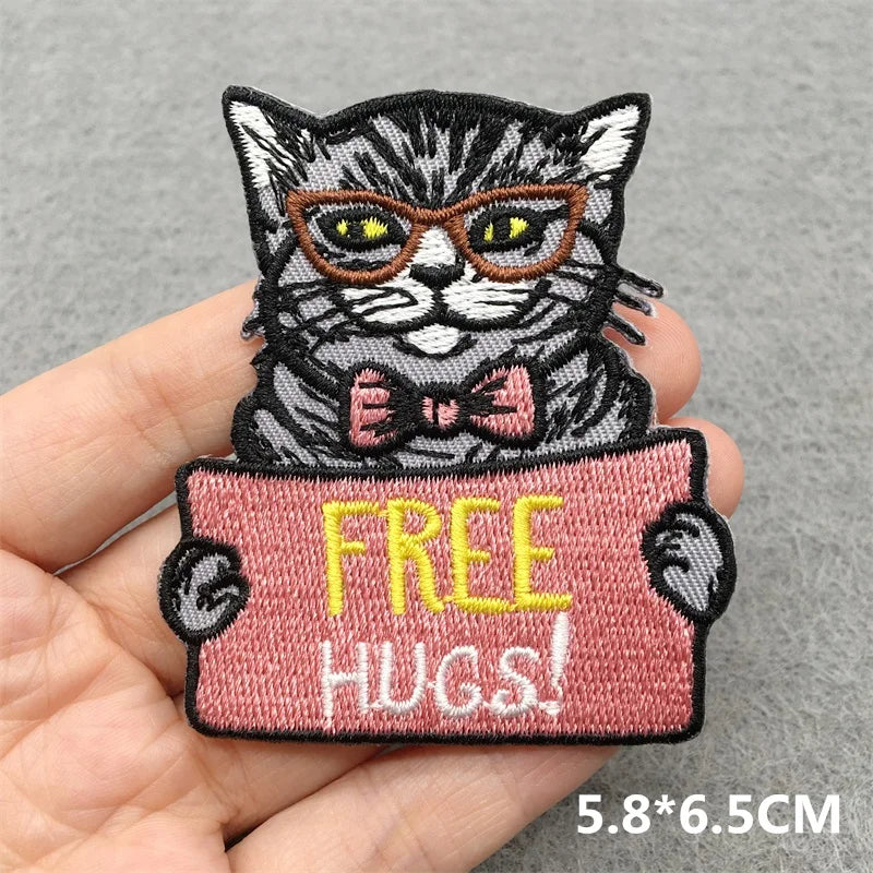 Embroidered patch of a cat holding a 'FREE HUGS!' sign held by a hand on a gray background.