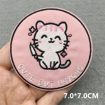 Circular patch with a cartoon cat and text 'Cute But Psycho' held in a hand against a gray background.