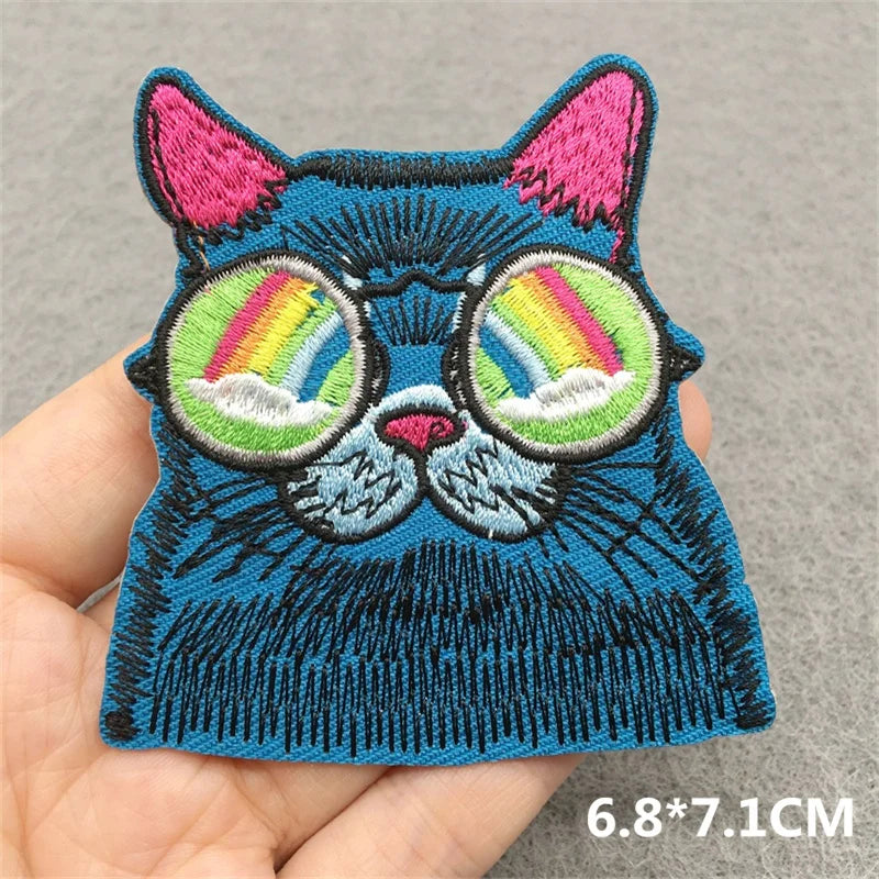 Embroidered patch of a blue cat with rainbow glasses held in a hand on a gray background.