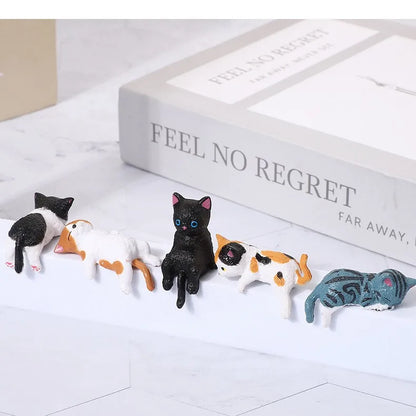 Small figurines of cats lined up on a white surface with a book titled 'Feel No Regret' in the background.