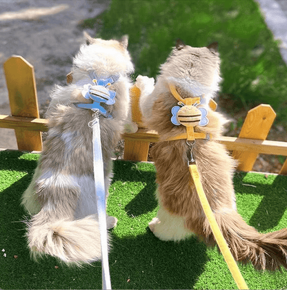 Two cat harnesses with a bee-themed design, one in blue and one in yellow, displayed on mannequin cats placed against a wooden fence and green moss background.