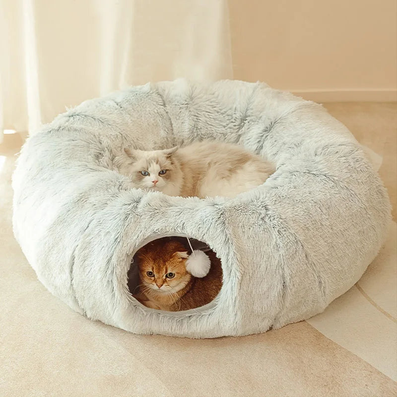 Two cats in a fluffy donut-shaped pet bed on a light-colored floor.