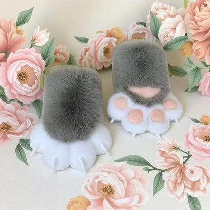 Two plush cat paw booties in gray and white faux fur, decorated with pink paw pads, displayed on a cream floral background.