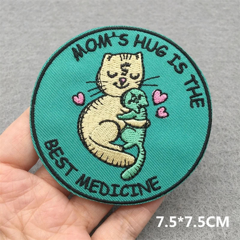 Circular patch with a cat holding a baby and text 'Mom's hug is the best medicine' held by a hand on a gray background.