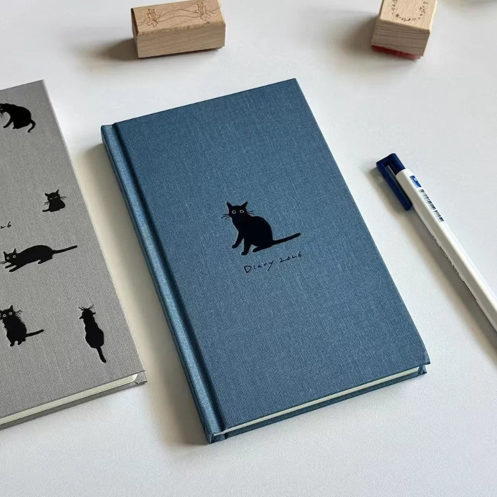 Blue notebook with black cat illustration on a white surface with stationery items.