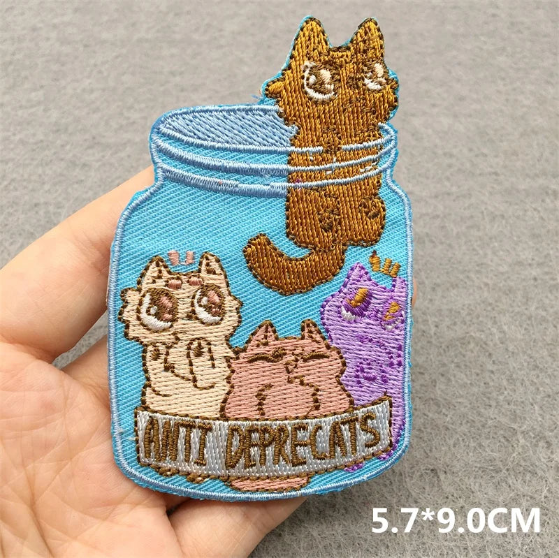 Embroidered patch with cats in a jar labeled 'Anti Deprecats' held by a hand on a gray background.