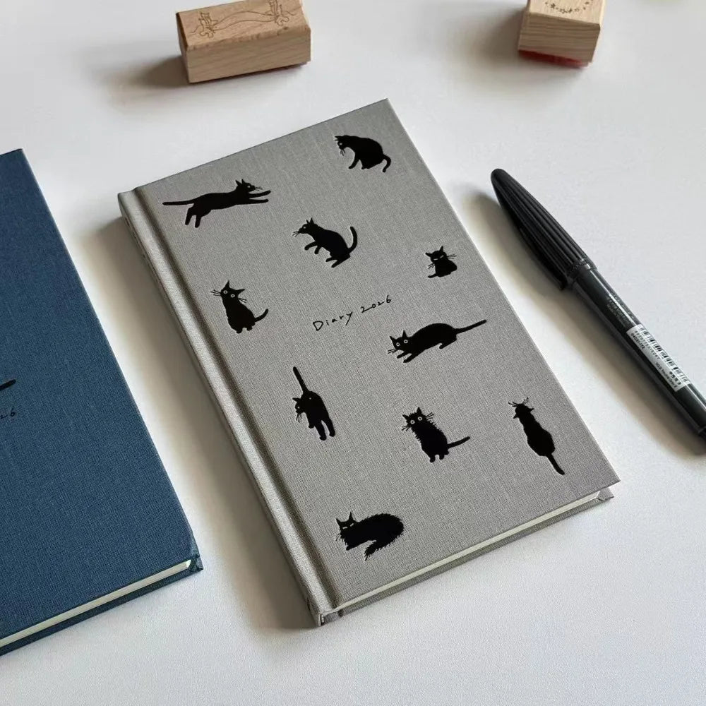 Grey notebook with black cat illustrations on a white surface with a pen and blue book.