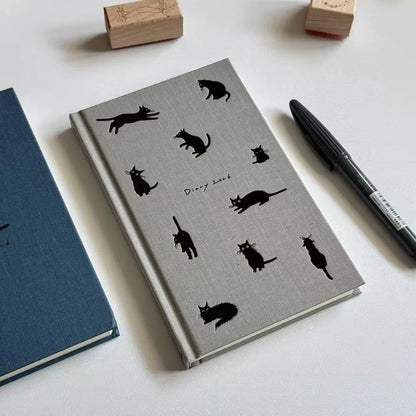 Grey notebook with black cat illustrations on a white surface with a pen and blue book.
