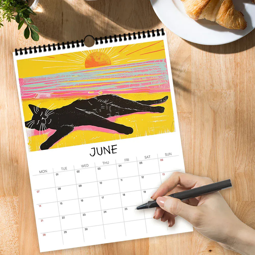 Calendar for June with a black cat illustration on a wooden table, hand holding a pen.