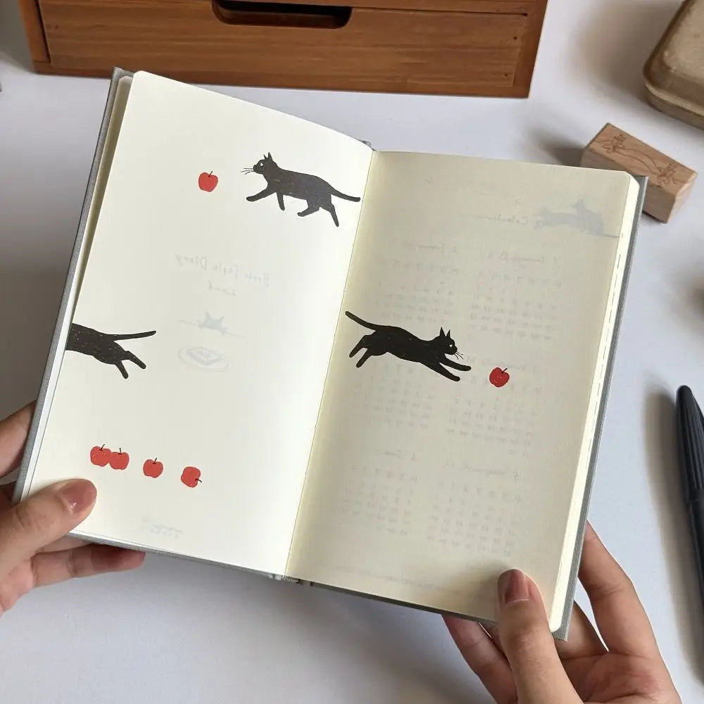 Open notebook with black cat illustrations and red apples on a white surface