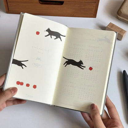 Open notebook with black cat illustrations and red apples on a white surface