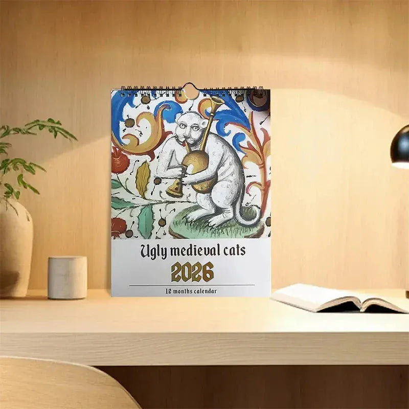 Calendar titled 'Ugly medieval cats 2026' on a wooden surface with a plant and lamp in the background.